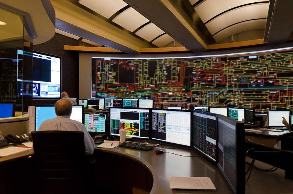 A control room operator sits at a round desk in front of several monitors.