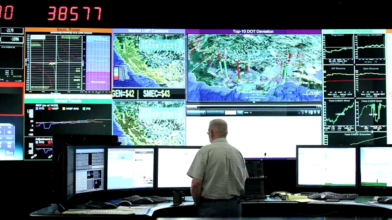 An operator standing in front of a computer and video wall analyzing data