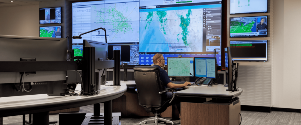 A controller sitting in a control room with a computer and a video wall