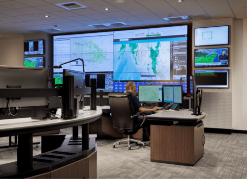 A controller sitting in a control room with a computer and a video wall